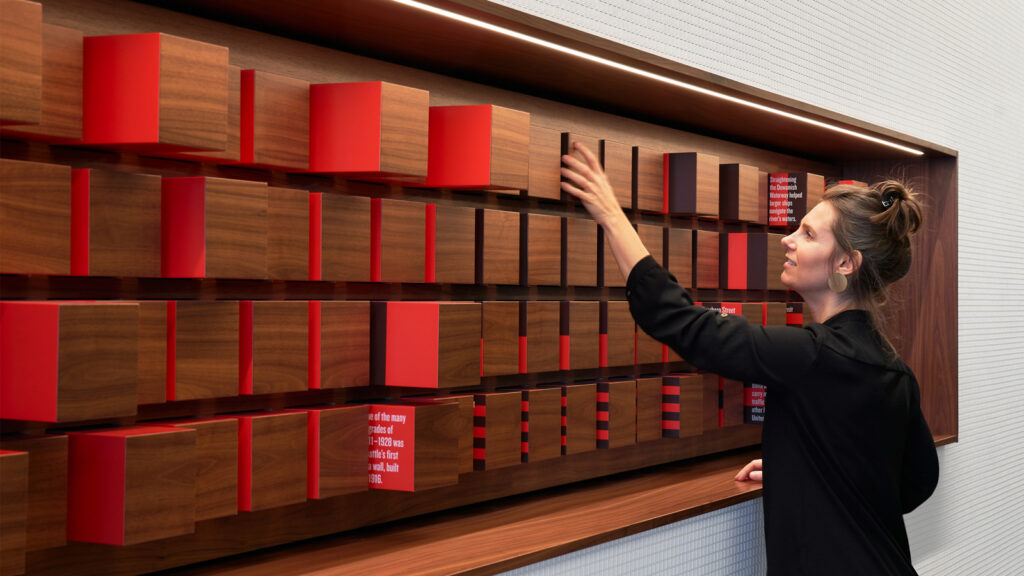 Person interacting with wooden wall graphic. Graphic has red cubes that can be pulled out of their slot to reveal information on the sides of the cubes